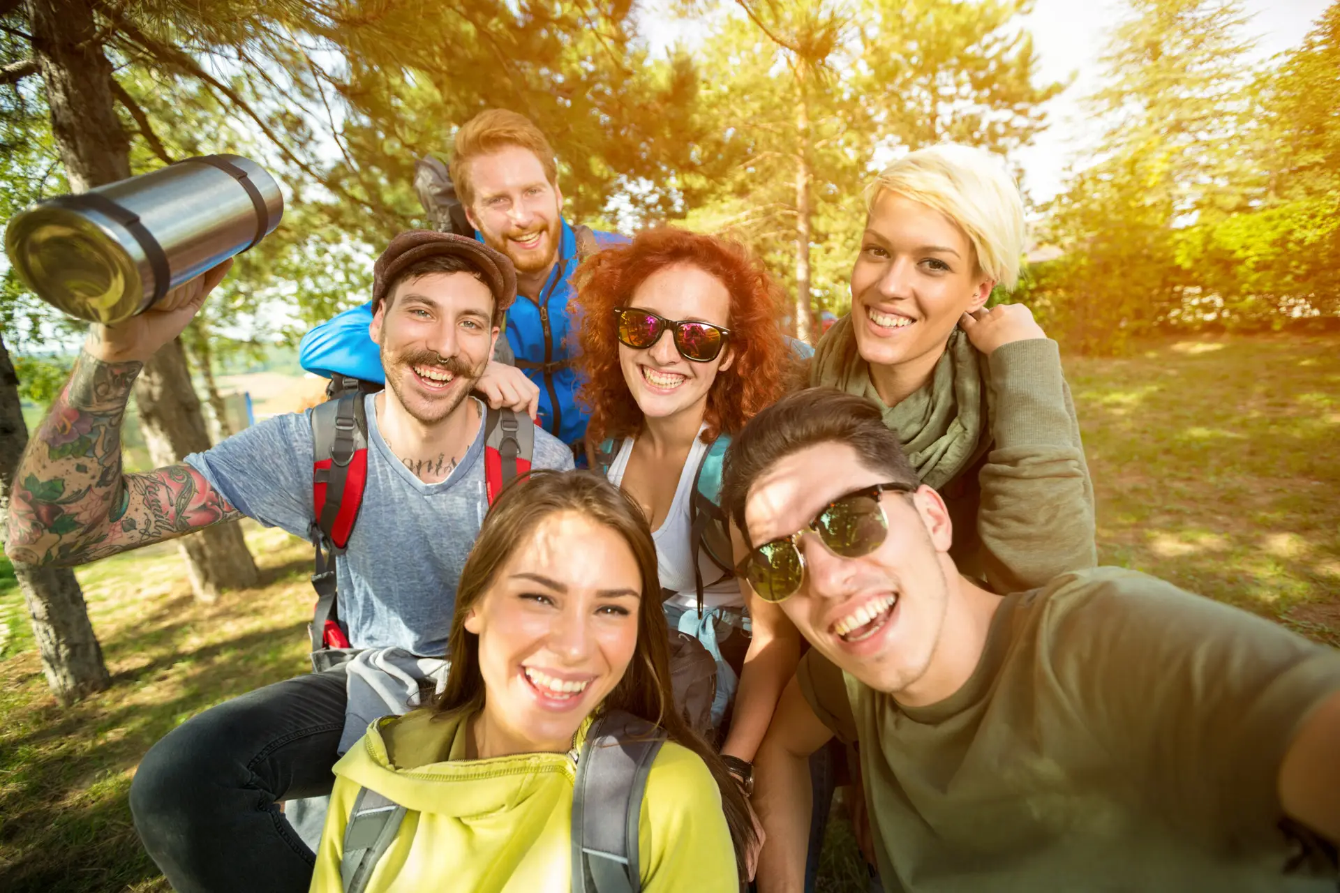 Eine fröhliche, diverse Gruppe von Freunden auf einer Wandertour, die in einem sonnendurchfluteten Wald lachend ein Selfie macht und dabei Gemeinschaft und Freude an Outdoor-Aktivitäten demonstriert.